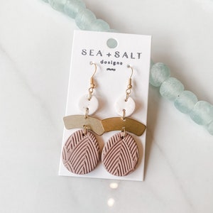 Könnte beinhalten: Ein Paar Ohrringe mit einem rosafarbenen Tonkreis mit geometrischem Muster, einer goldenen Stange und einem weißen Keramikkreis. Die Ohrringe befinden sich auf einer weißen Karte mit dem Text "SEA + SALT designs".