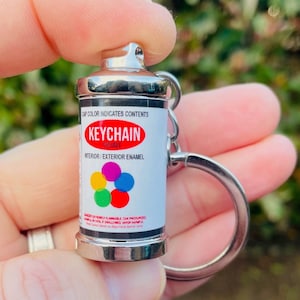 Könnte beinhalten: Ein silberner Mini-Sprühdosen-Schlüsselanhänger mit weißem Etikett. Auf dem Etikett steht "KEYCHAIN PAINT" in Rot, darunter "INTERIOR / EXTERIOR ENAMEL". Die Dose hat ein farbenfrohes Punktdesign und einen silbernen Schlüsselring.