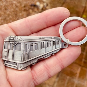Może przedstawiać: Srebrny brelok do kluczy w kształcie pociągu metra. Brelok przedstawia szczegółową miniaturową replikę wagonu metra, z oknami, drzwiami i kołami. Pociąg jest przymocowany do srebrnego kółka na klucze.