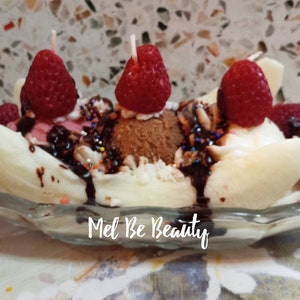 May include: A banana split candle with three red raspberries, chocolate sauce, and sprinkles. The candle is in a clear glass dish and has the text "Mel Be Beauty" on the bottom.