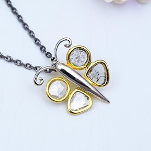 May include: A silver butterfly pendant necklace with a gold and diamond-like stone design. The butterfly has four stones and a silver chain. The necklace is displayed on a white surface with flowers.