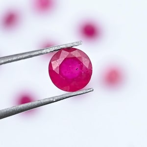 Peut inclure: Un rubis rond et facetté, d'un rouge profond, est maintenu par une pince argentée sur fond blanc. Plusieurs autres rubis flous sont dispersés en arrière-plan. La pierre précieuse est d'une couleur vive et saturée.