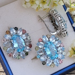 Puede incluir: Pendientes de plata con diseño floral. Cada pendiente presenta una piedra preciosa grande, ovalada, de color azul claro, rodeada de piedras preciosas más pequeñas y multicolores. Los pendientes se exhiben en una caja azul con flores blancas de fondo.