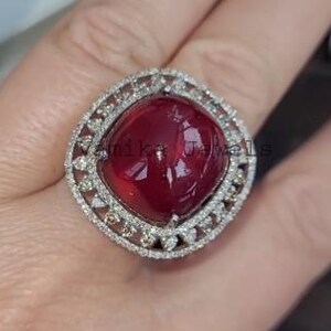 Può includere: Un anello grande, di forma quadrata, con una vibrante pietra preziosa rosso intenso al centro. La pietra preziosa è circondata da un alone di piccoli diamanti scintillanti, incastonati in una fascia di metallo argentato. L'anello è mostrato su una mano.