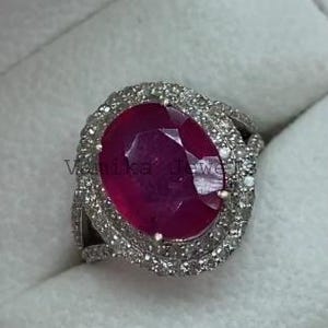 May include: A silver ring featuring a large, oval-cut ruby gemstone. The ruby is a deep red color and is surrounded by a halo of smaller, clear gemstones. The ring is set in a white box.
