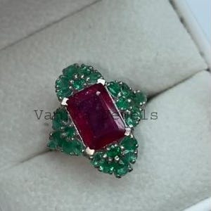 May include: A gemstone ring featuring a large, rectangular ruby center stone. The ruby is surrounded by smaller, round emeralds in a floral design. The ring is set in a gold band and is displayed in a white jewelry box.