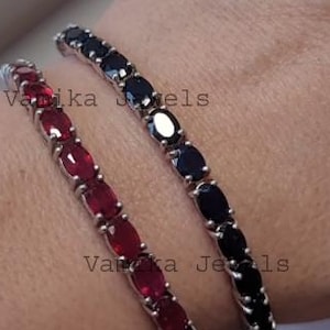 Può includere: Due braccialetti con pietre preziose, uno con pietre rosse e l'altro con pietre nere, sono esposti su un polso. I braccialetti hanno una montatura color argento e sono adornati con pietre preziose taglio ovale. I braccialetti sono di Vanika Jewels.