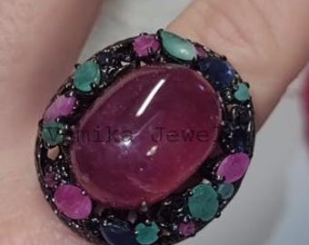May include: An eye-catching ring with a large, oval, ruby-red central stone. The ring is encircled by smaller gemstones in green, pink, and blue. The dark band of the ring has the text "mika Jewel". A striking piece of jewellery.