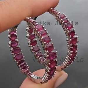 May include: A pair of silver hoop earrings, each lined with oval-cut, deep red gemstones. The earrings are held up against a blurred background of water and greenery, showcasing the jewelry's sparkle and design.