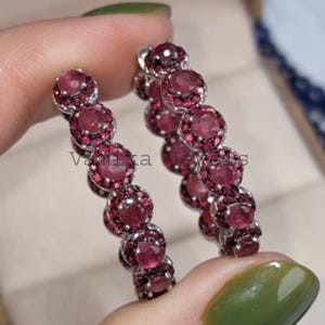 May include: A pair of hoop earrings with a series of round, deep red gemstones set in a silver-toned metal. The earrings are held in front of a tan jewelry box. The gemstones are a vibrant, rich color.