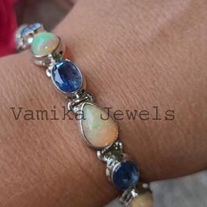 May include: A silver bracelet with alternating teardrop-shaped opal and blue gemstone beads. The bracelet is on a person's wrist and the text "Vamika Jewels" is visible.