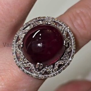 Può includere: Un anello grande con una pietra preziosa rosso scuro, probabilmente un rubino. La pietra centrale, tagliata a cabochon, è circondata da un alone di piccoli diamanti trasparenti incastonati in una montatura in argento o oro bianco. L'anello è su un dito.