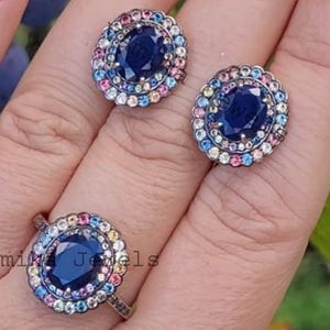 May include: A set of blue sapphire earrings and a ring with a halo of colorful gemstones. The ring is set in silver and has a Vami Jewels logo on the band.