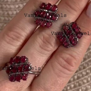 Puede incluir: Un conjunto de joyas con un anillo y dos pendientes. Cada pieza tiene un grupo de pequeñas piedras preciosas ovaladas, de color rojo intenso, engastadas en un metal oscuro. El anillo está en un dedo y los pendientes en una mano. El texto "Vam Jewels" es visible.