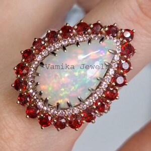 May include: A large, pear-shaped ring featuring a central opal stone with iridescent colors. The opal is surrounded by a halo of small, red gemstones and a ring of smaller, pink stones. The ring is set in a gold-colored metal.