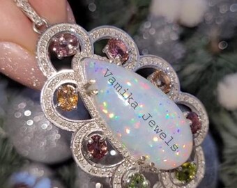 May include: A silver pendant featuring a large, iridescent opal. The opal is set within a floral design, accented with small, colourful gemstones and a halo of clear stones. The text "Vamika Jewels" is visible on the opal.