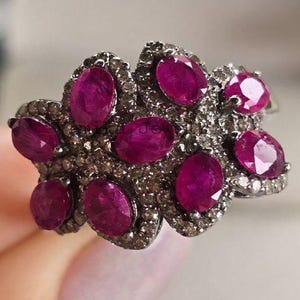 May include: A cluster ring with oval rubies set in a dark metal, surrounded by smaller stones. The ring has a floral design, with multiple vibrant red gemstones.