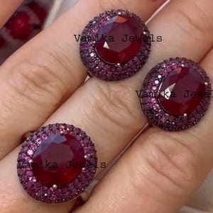 May include: A set of jewelry featuring a ring and two earrings. Each piece has a large, oval-shaped, deep red gemstone surrounded by smaller, dark pink stones. The text "Vanika Jewels" is visible on each piece.