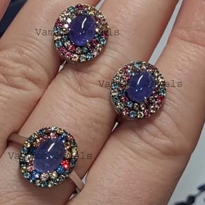 May include: Three rings featuring a central oval, deep blue gemstone encircled by a halo of smaller, colourful gemstones. The rings are set on a silver band. The image is well-lit, highlighting the jewellery's details.