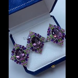 May include: A set of three amethyst rings displayed in a blue jewelry box. Each ring features a large, oval-cut amethyst surrounded by smaller purple and green gemstones. The rings are set on a white interior.