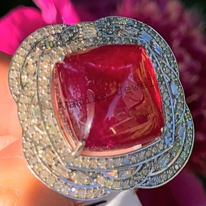 May include: A silver ring featuring a large, square, red gemstone surrounded by smaller diamonds. The ring is set in a delicate, intricate design.