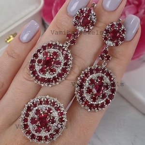 May include: A jewelry set featuring a ring and a pair of earrings. The earrings and ring have a circular design with a central oval-shaped ruby surrounded by smaller rubies and clear stones. The text "Vamika Jewels" is visible.