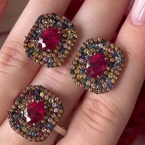 以下が含まれることがあります： 正方形のデザインの3つのリング。各リングの中央には、大きな楕円形の濃い赤色の宝石があり、その周りを小さなカラフルな宝石のモザイクが囲んでいます。リングはシルバーのバンドにセットされています。「Yamika Jewels」の文字が見えます。