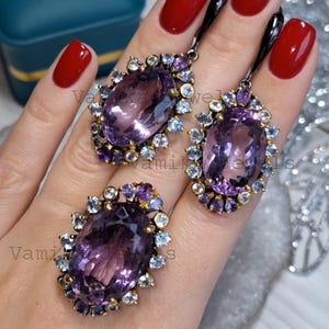 Peut inclure: Un ensemble de bijoux comprenant une bague en améthyste ovale et de grandes boucles d'oreilles assorties. Les pierres d'améthyste sont entourées de petites pierres précieuses violettes et bleu clair, serties dans un métal doré. Les boucles d'oreilles ont de longs crochets noirs.