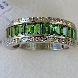 Op de afbeelding: Een close-up van een zilveren ring met een rij rechthoekige, heldergroene edelstenen in het midden, omlijst door twee rijen kleine, heldere edelstenen. De ring heeft de tekst "Vamika Jewels" erop.