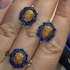 May include: Three silver rings with octagonal settings featuring a large oval opal surrounded by a halo of smaller blue sapphires.