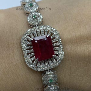 May include: A silver bracelet featuring a large, rectangular, deep red gemstone at its center, surrounded by a halo of smaller, clear stones. The bracelet has a repeating pattern of circular links with green and clear stones. The text "Vamika Jewels" is visible.