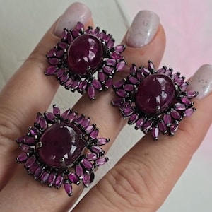 以下が含まれることがあります： リングとイヤリング2つの3点セットのジュエリー。各アイテムは、小さなマゼンタ色の花びら型の石に囲まれた、大きな楕円形の濃い赤色の宝石を特徴としています。金属のセッティングは濃い灰色です。