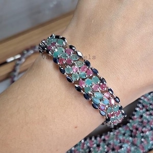 Puede incluir: Un brazalete de piedras preciosas multicolor con gemas de corte ovalado en tonos turquesa, rosa y azul oscuro. El brazalete está engastado en metal plateado y se muestra en una muñeca.