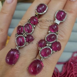 Puede incluir: Un par de pendientes colgantes con piedras preciosas rosas oscuras ovaladas y redondas. Las piedras preciosas están engastadas en un metal plateado con un halo de pequeñas piedras transparentes. Los pendientes se sostienen en una mano.