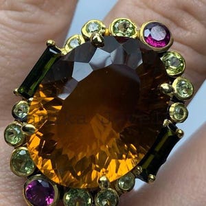 Könnte beinhalten: Ein goldfarbener Ring mit einem großen, facettierten, bernsteinfarbenen Edelstein als Mittelpunkt. Der Ring ist mit kleineren, runden, hellgrünen und tiefmagentafarbenen Edelsteinen sowie dunkelgrünen rechteckigen Steinen besetzt.