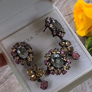 May include: A pair of dangle earrings with a light blue gemstone surrounded by smaller clear, yellow, and pink gemstones. The earrings are in a white box with the text "Mika Jewel" on the box.