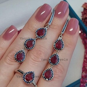Puede incluir: Un conjunto de joyas que incluye un anillo y pendientes. Los pendientes y el anillo tienen un diseño de lágrima con una gran piedra preciosa roja rodeada de pequeñas piedras preciosas azules. La joyería es de metal plateado.