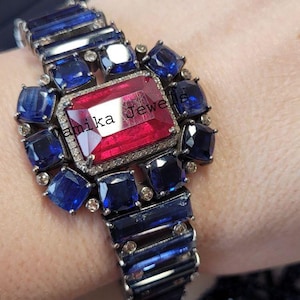 May include: A striking bracelet featuring a large, rectangular red gemstone at its center, surrounded by smaller, square blue gemstones. The bracelet has a dark metal setting and is adorned with the text "amika Jewe".