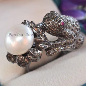 May include: A silver ring featuring a detailed frog design with a large white pearl. The frog's body is encrusted with small stones, and it has a single red gemstone eye. The ring is set against a soft, blurred background.