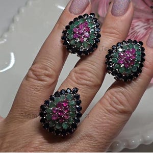 Op de afbeelding: Drie edelsteenringen met een druppelontwerp. Elke ring heeft een cluster van kleine, ronde edelstenen in zwart, groen en roze. De ringen zijn geplaatst op een lichtgekleurd oppervlak.