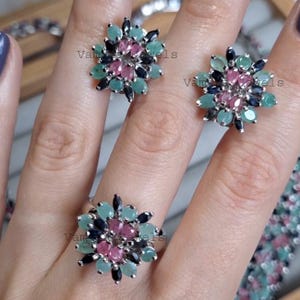 Könnte beinhalten: Ein silbernes Schmuckset mit Blumenmuster, bestehend aus einem Ring und zwei Ohrringen. Jedes Stück ist mit einer Ansammlung von rosa, türkis und dunkelblauen Edelsteinen besetzt, die wie Blütenblätter angeordnet sind.