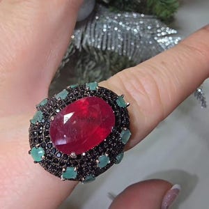 May include: A ring featuring a large, oval-cut, deep red central gemstone surrounded by a halo of small, round, light green stones and a dense layer of tiny black stones. The setting is dark metal, and the text "Vimaka Jewels" is visible.