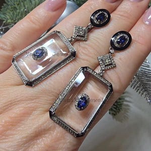 Pode incluir: Um par de brincos pendurados prateados. Cada brinco apresenta uma moldura retangular transparente com uma pedra preciosa azul central e uma borda de pequenas pedras claras. Os brincos têm tops ovais pretos com pedras azuis e detalhes em forma de diamante.