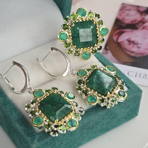 May include: A jewelry set featuring a ring and earrings with large, square-cut emerald-green stones. The ring and earrings are surrounded by smaller green and gold-colored stones, set in a silver-colored metal. The set is displayed in a teal-colored jewelry box.
