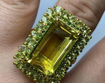 May include: A gold ring with a large, rectangular yellow gemstone, framed by a halo of smaller, light green gemstones. The ring's design is detailed, highlighting the vivid colours and the contrast between the yellow and green stones. A statement ring.