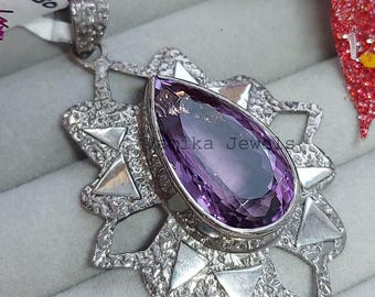 May include: A silver pendant with a large, pear-shaped purple gemstone at the center. The pendant has a starburst design with triangular accents. The background includes a tag and decorative elements.