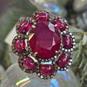 Peut inclure: Une bague en forme de fleur avec un grand rubis ovale au centre, entouré de petits rubis et d'un halo de petites pierres précieuses claires. La bague a un anneau de couleur argent.