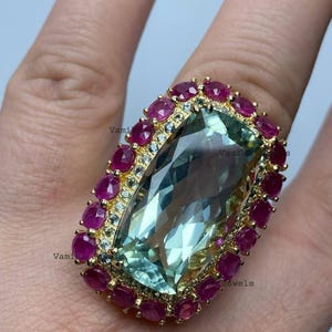Puede incluir: Un anillo dorado con una gran gema rectangular verde claro en el centro, rodeada por un borde de pequeñas gemas rojas redondas. El diseño del anillo incluye un engaste delicado con piedras claras más pequeñas.