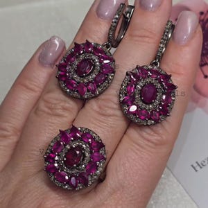 May include: A jewellery set comprising a ring and earrings. Each item features a central oval ruby encircled by smaller, teardrop-shaped rubies. The setting is a dark metal with small, clear stones around the central ruby.