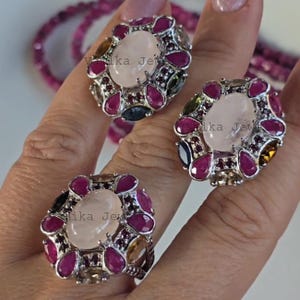 Könnte beinhalten: Drei verzierte Ringe mit einem zentralen ovalen, hellrosa Stein, umgeben von kleineren, tropfenförmigen, tiefroten und andersfarbigen Edelsteinen. Die Ringe sind auf einer Hand platziert, mit einer passenden Perlenkette im Hintergrund.